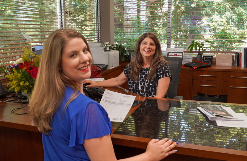 Two women at a desk in a small business office with large windows.