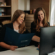 One woman shows another woman something on her computer screen while holding a folder.