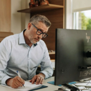 A man sitting at a desk writing down notes from his computer.