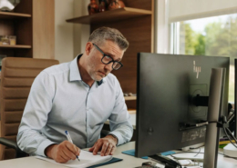 A man sitting at a desk writing down notes from his computer.