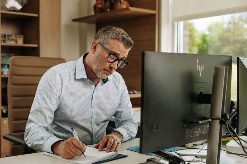 A man sitting at a desk writing down notes from his computer.
