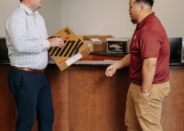 Two men are talking while one is holding a laptop package.
