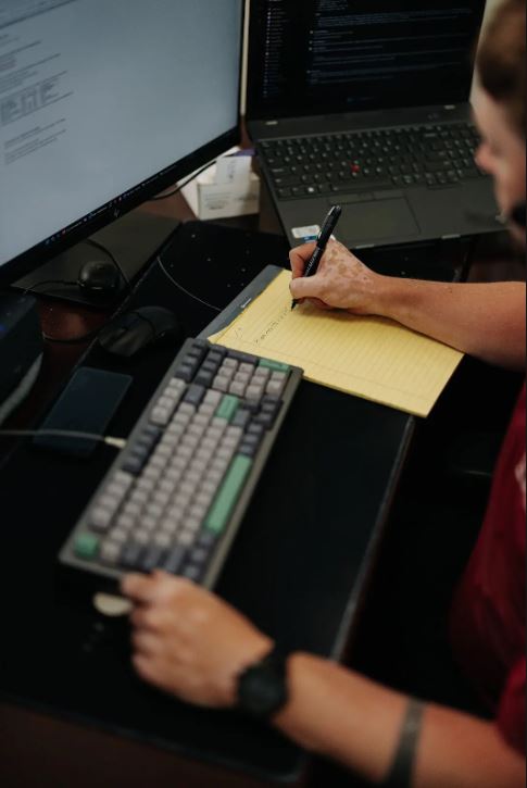 An IT support person writing on a notepad next to a computer keyboard.