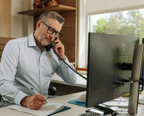 A remote office worker talks on the phone in front of his computer at his desk.