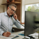 A remote office worker talks on the phone in front of his computer at his desk.